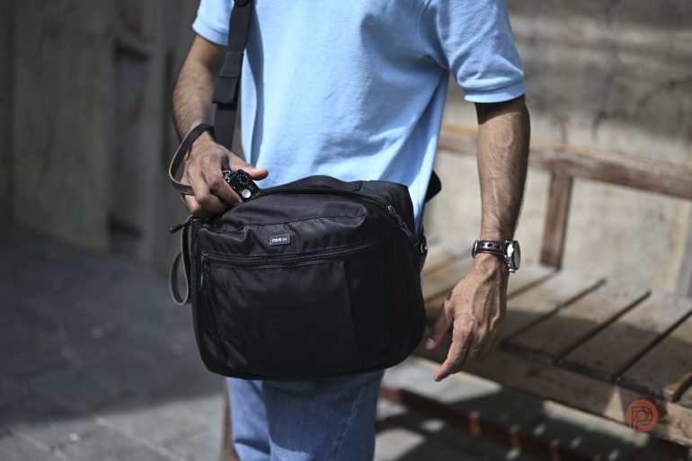 Man in a light blue shirt holds a black shoulder bag and camera, standing next to a wooden bench.