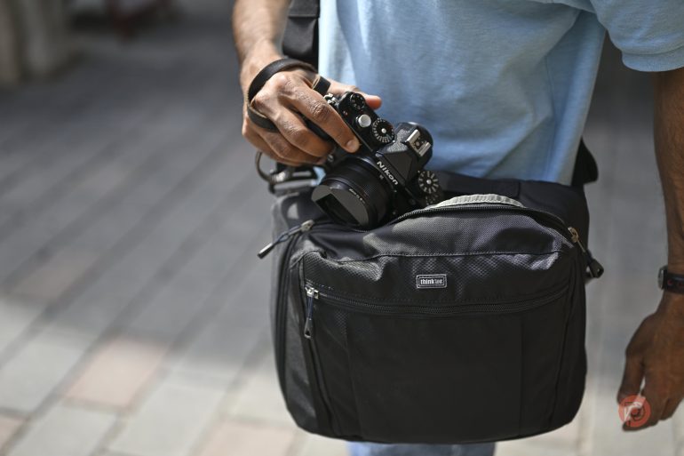 Person in blue shirt puts a black camera into a black shoulder camera bag outdoors.