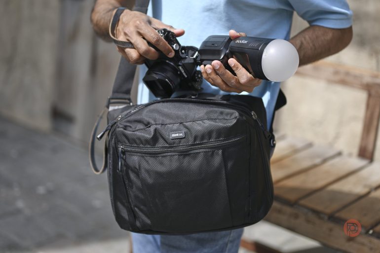 A person wearing a blue shirt holds a camera with a flash and carries a black shoulder bag.