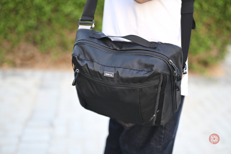 Person carrying a black messenger bag with a shoulder strap, standing on a paved path outdoors.
