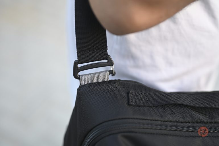 Close-up of a black bag strap attached to a shoulder bag, worn by a person in a white shirt.