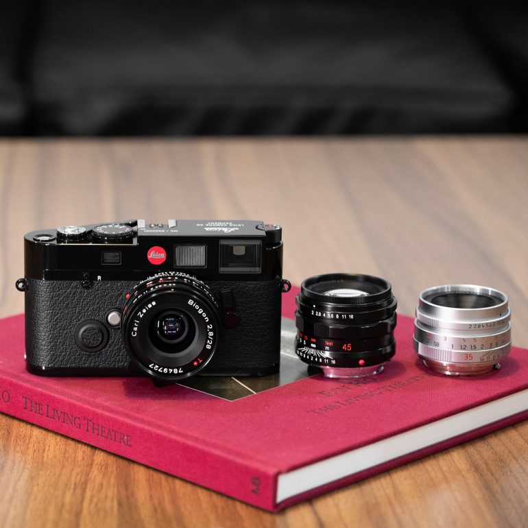 A Leica camera and two lenses rest on a red book titled The Living Theatre on a wooden surface.