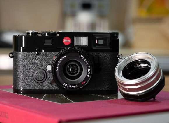 A black Leica camera with a Zeiss lens sits on a red book, next to a detached camera lens.