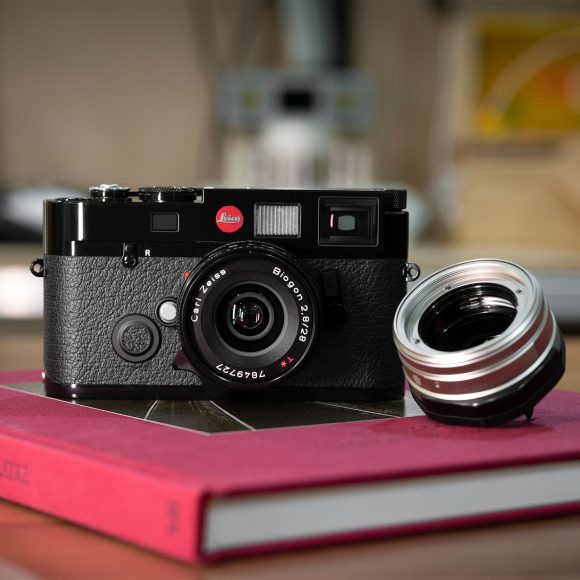 A black Leica camera with a Zeiss lens sits on a red book, next to a detached camera lens.