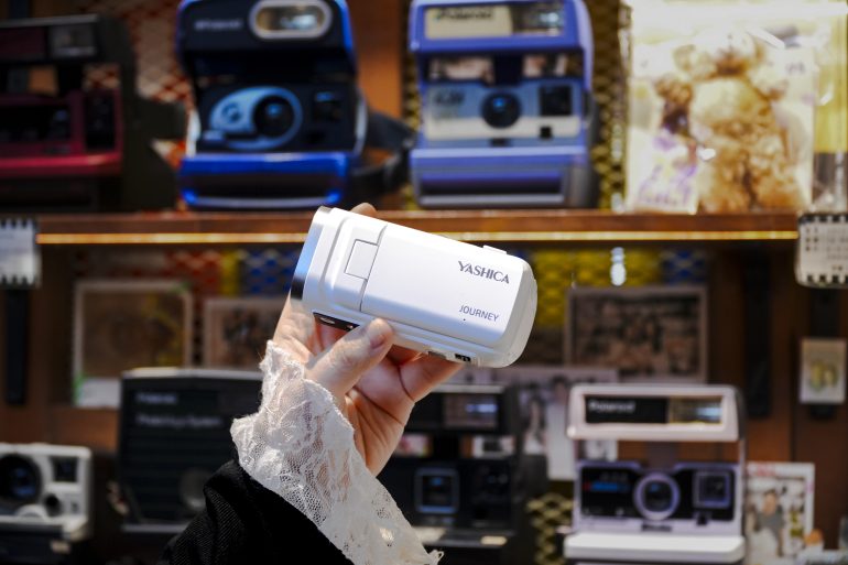 A hand holds a white Yashica Journey camera in front of a display of vintage cameras on shelves.