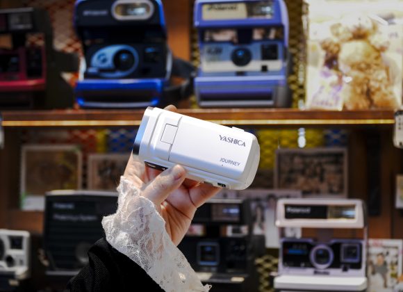 A hand holding a white Yashica camera in front of a display of vintage cameras and photos.