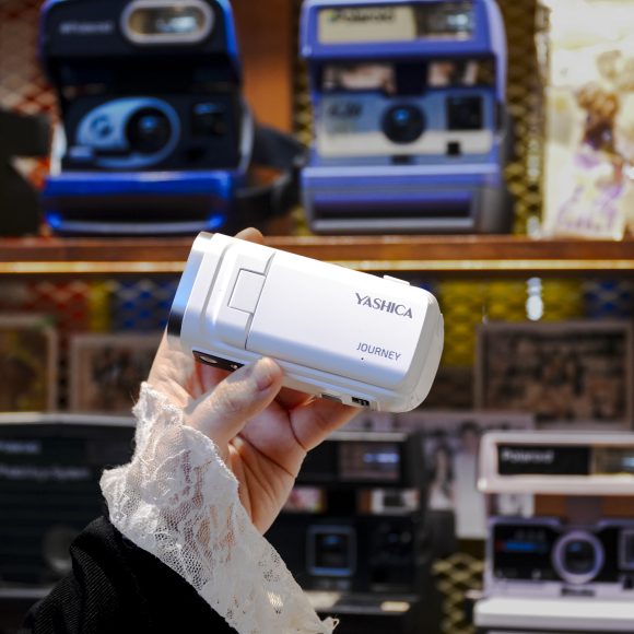 A hand holding a white Yashica camera in front of a display of vintage cameras and photos.