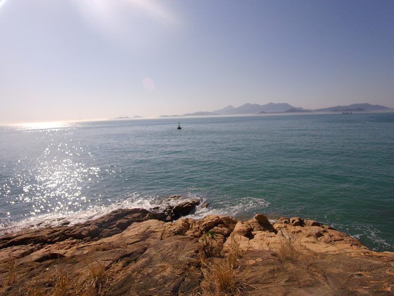 Rocky coastline with gentle waves, sparkling blue sea, and distant mountains under a clear, sunny sky.
