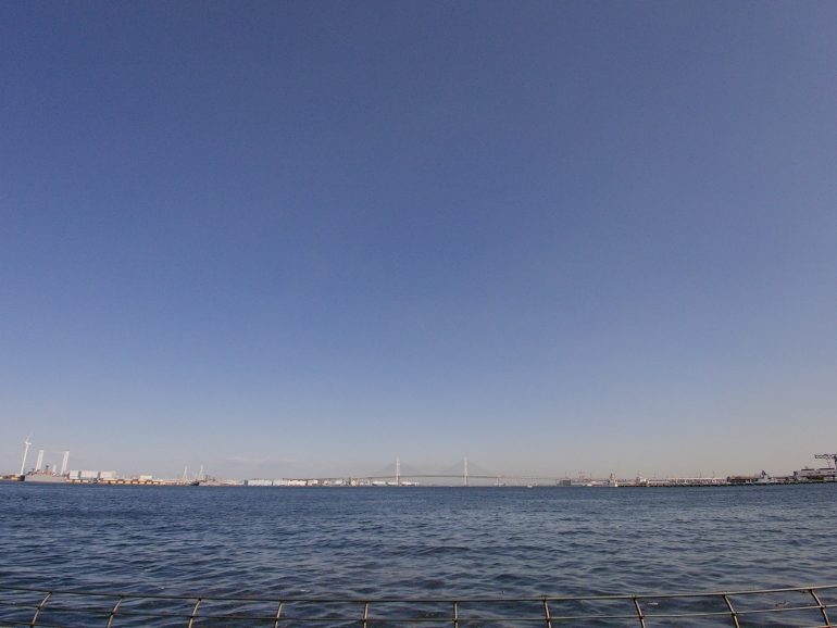 A calm harbor with distant cranes and wind turbines under a clear blue sky.