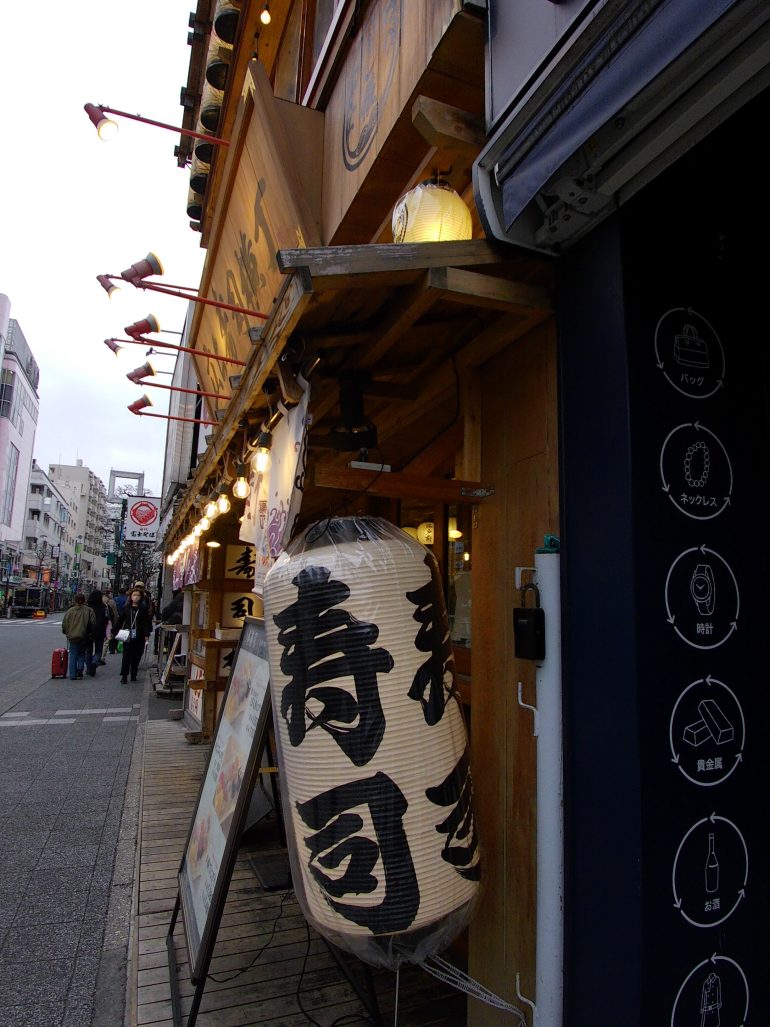 Large paper lantern with Japanese characters outside a sushi restaurant on a city street in Japan.