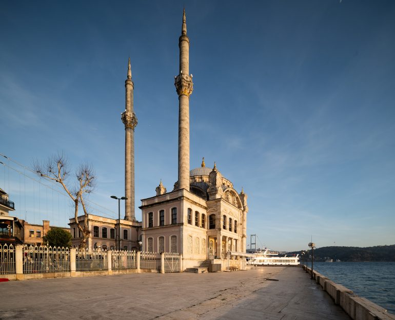 A mosque with two tall minarets stands by the waterfront under a clear blue sky.