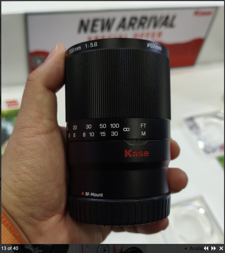 A hand holding a Kase RF-mount camera lens in front of a New Arrival display sign.