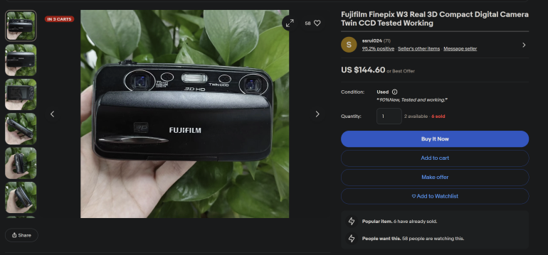 A hand holds a Fujifilm Finepix W3 3D camera in front of green leafy plants, shown on an eBay listing page.