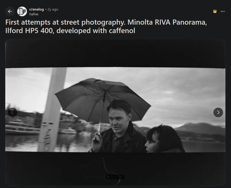 A man and a woman walk under an umbrella on a street, with water and hills in the background. Black and white photo.