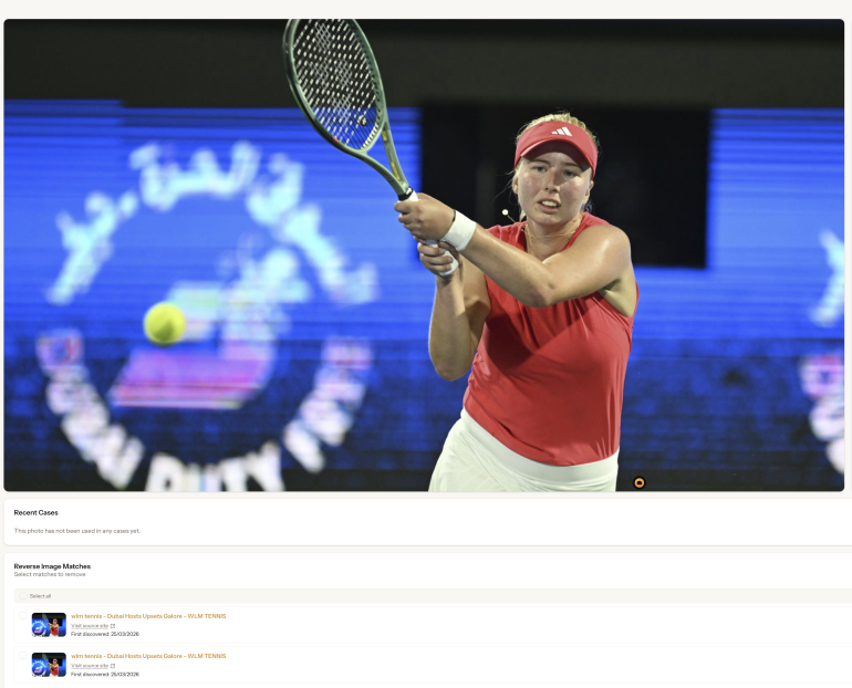A tennis player in a red outfit hits a backhand shot during a match on an outdoor court at night.