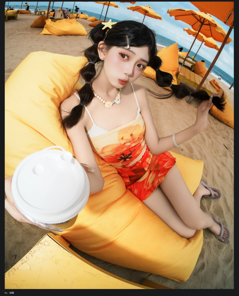 Young woman in a floral dress sits on a yellow beanbag at the beach, holding a drink and playing with her braids.