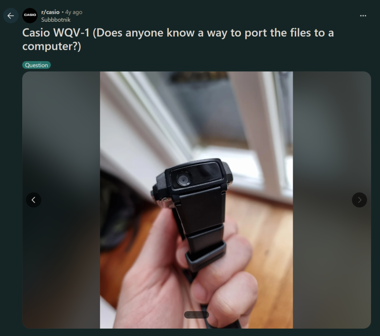 A hand holding a Casio WQV-1 watch with a camera near a window on a wooden floor.