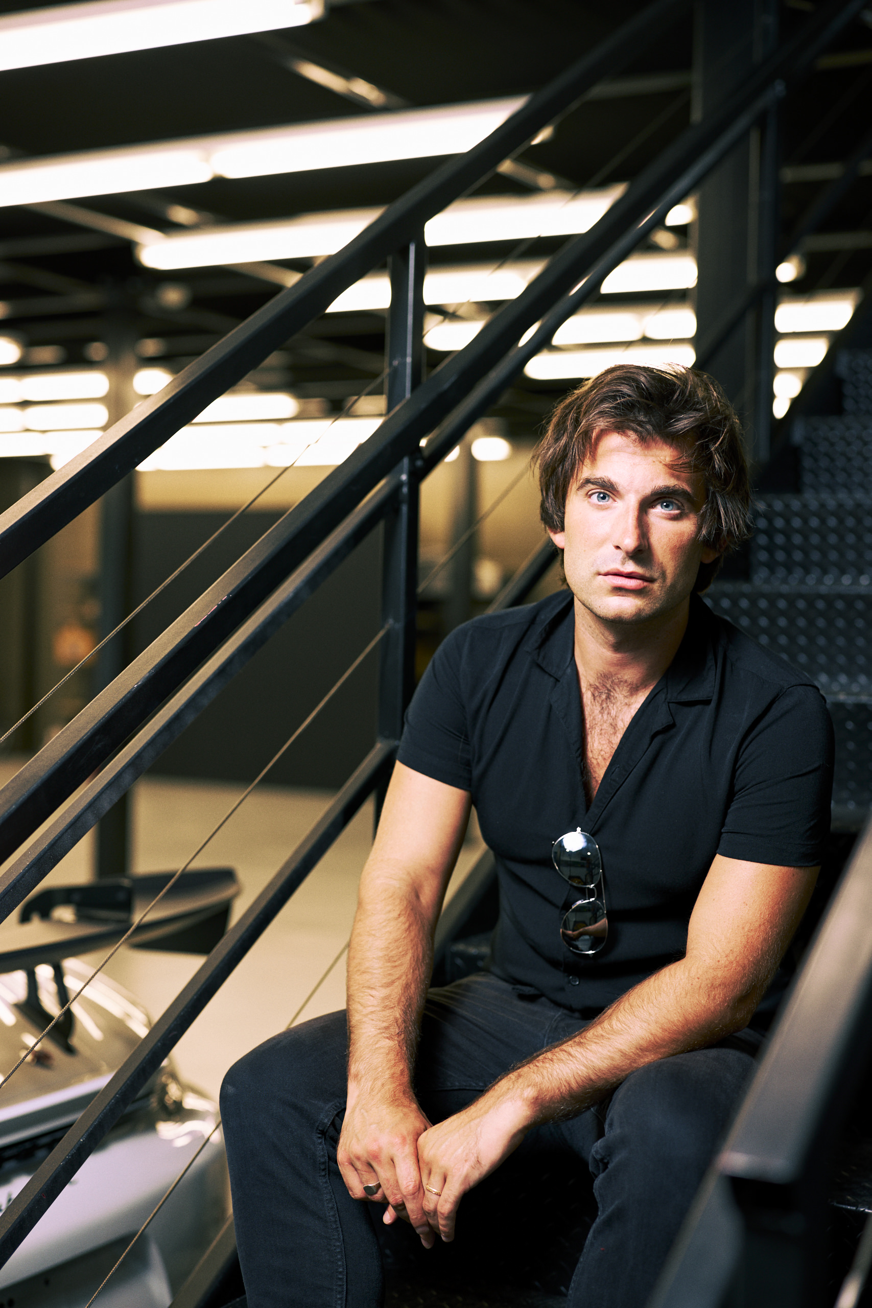 A man in a black shirt sits on a staircase, looking at the camera, with a car in the background.