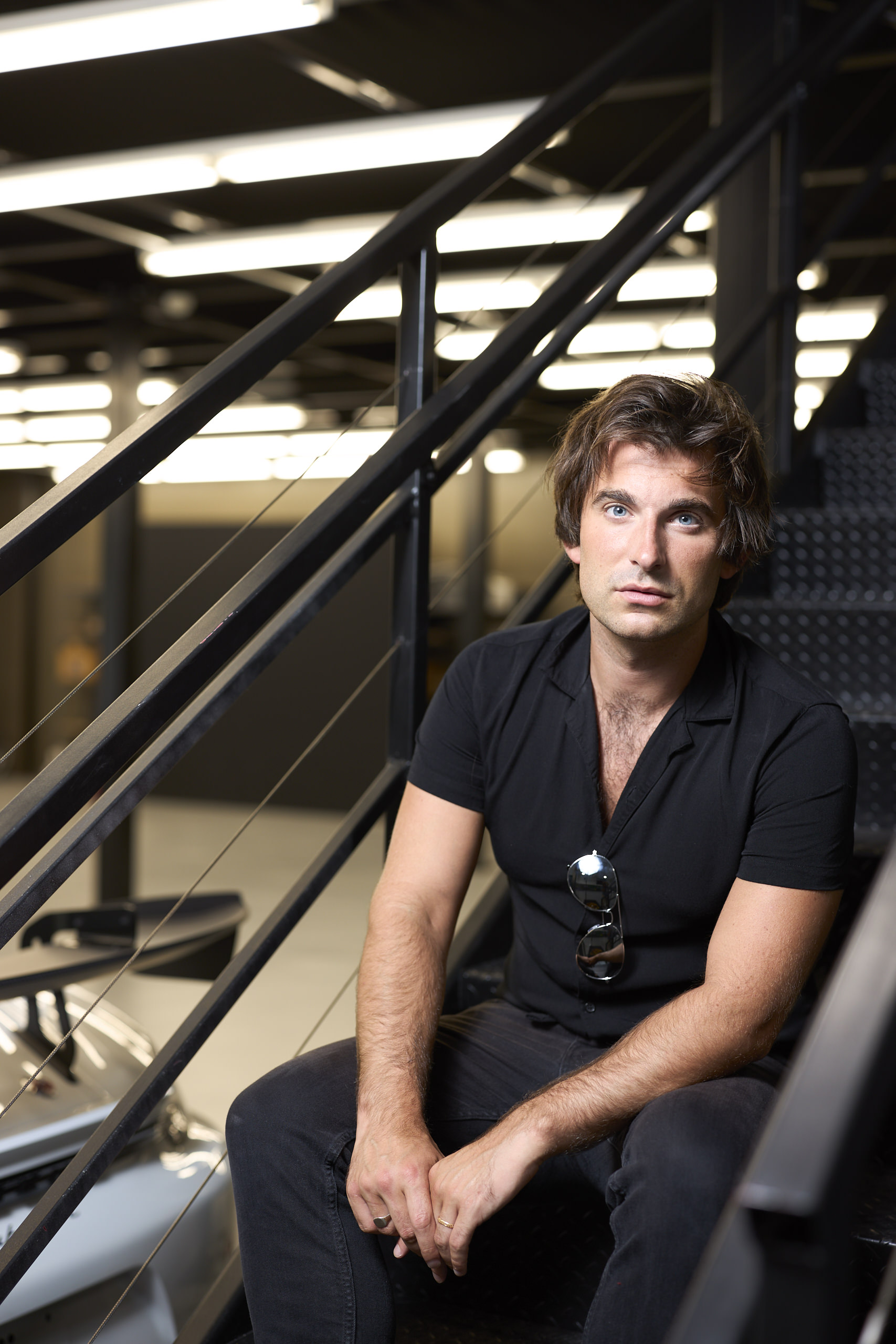 A man in a black shirt sits on metal stairs indoors, with a car partially visible beside him.
