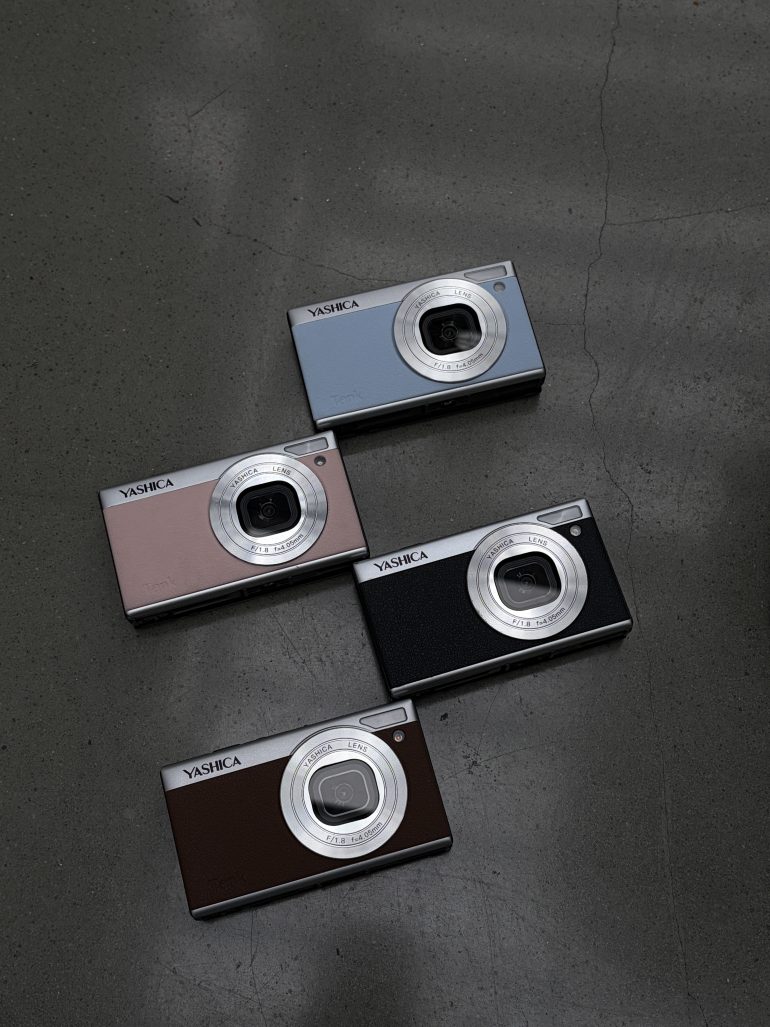 Four Yashica digital cameras in different colors are arranged on a gray, cracked concrete surface.