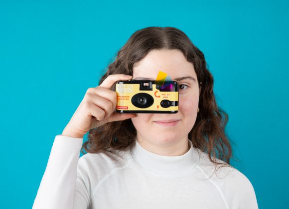 A woman in a white shirt holds a yellow camera up to her eye against a blue background.