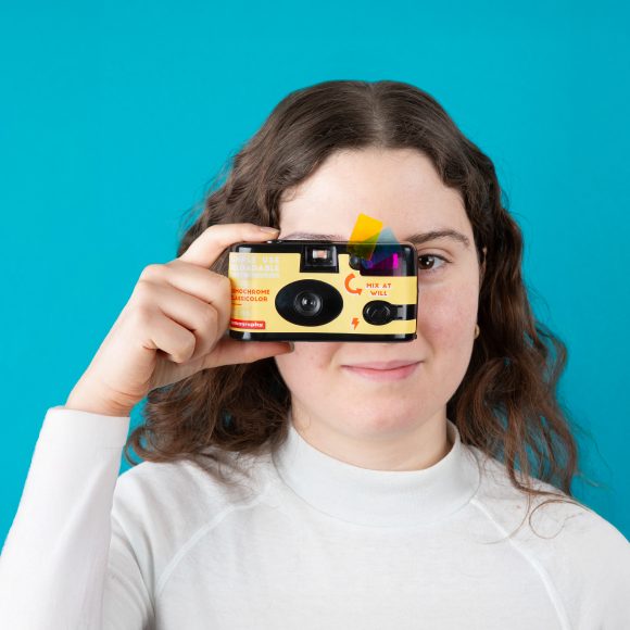 A woman in a white shirt holds a yellow camera up to her eye against a blue background.