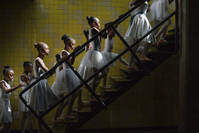 A group of young ballerinas in white tutus ascend a staircase against a yellow tiled wall.