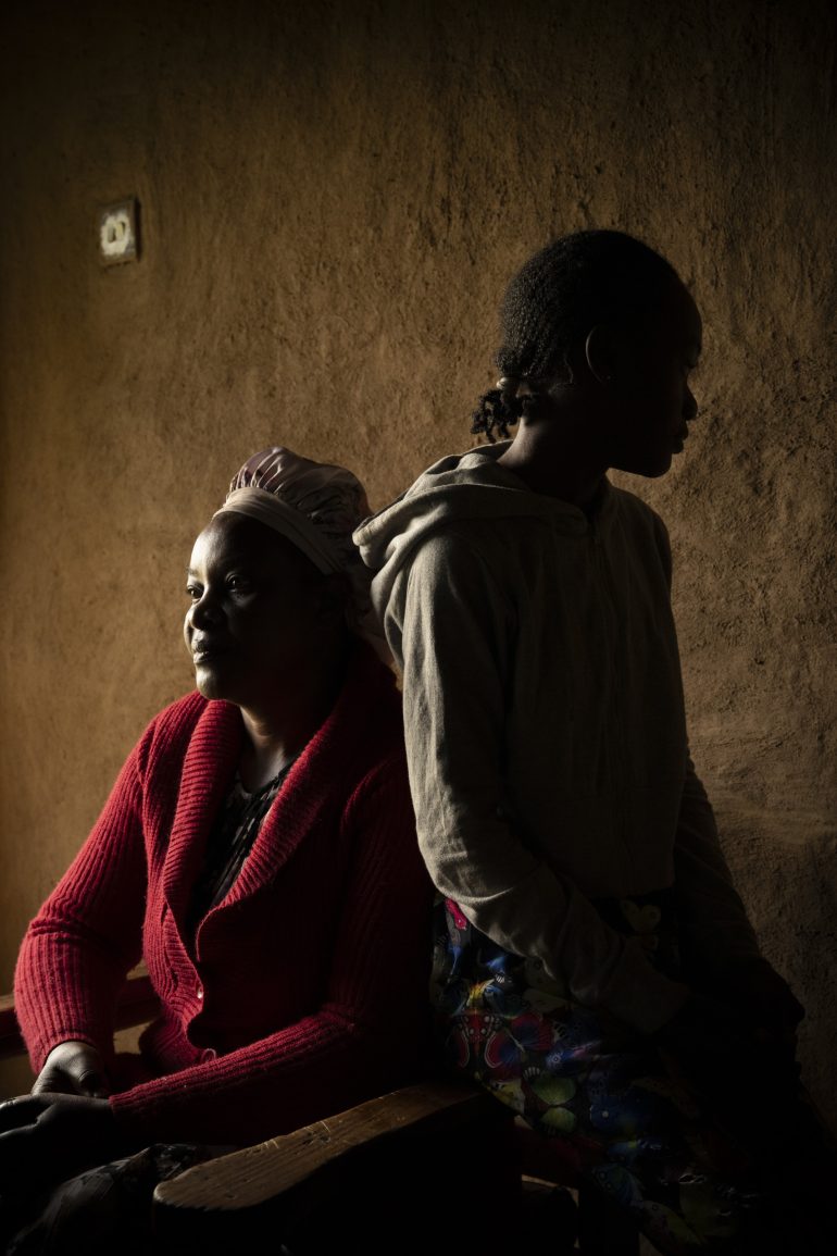 Two people sit side by side in low light, one facing forward and the other in profile against a textured wall.
