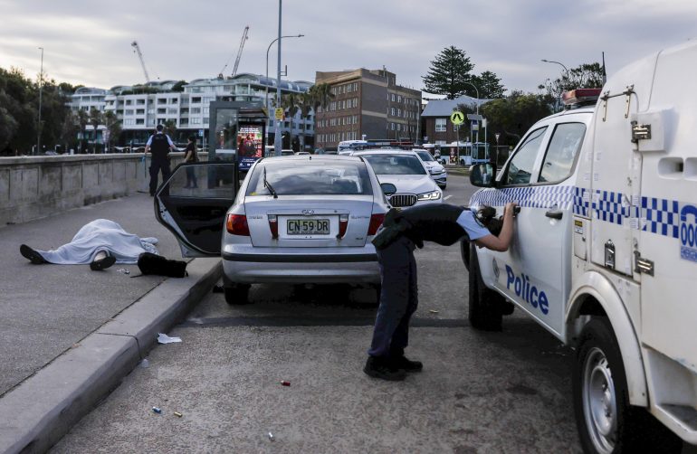 A police officer leans on a police vehicle near a crashed car and a person lying on the ground.