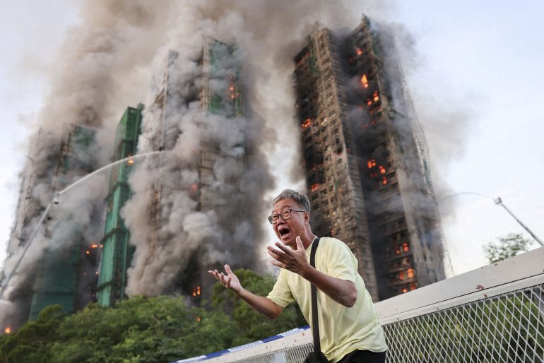 A distressed man gestures in front of burning high-rise buildings with heavy smoke and flames.