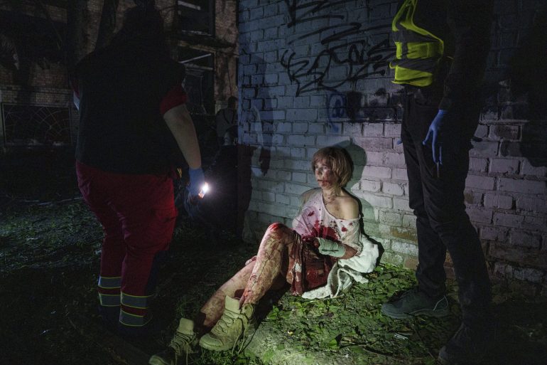 A bloodied woman sits against a graffiti wall at night, surrounded by emergency responders with flashlights.