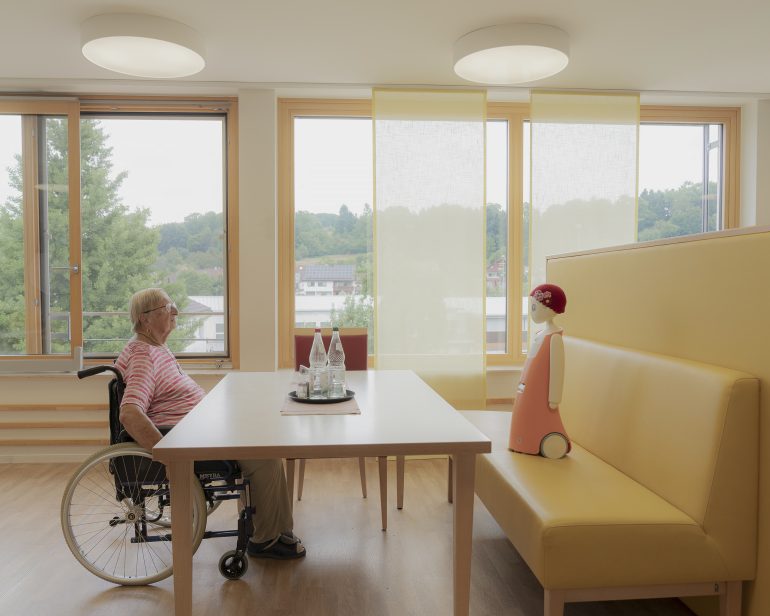 An elderly person in a wheelchair faces a robot across a table in a bright, modern room with large windows.