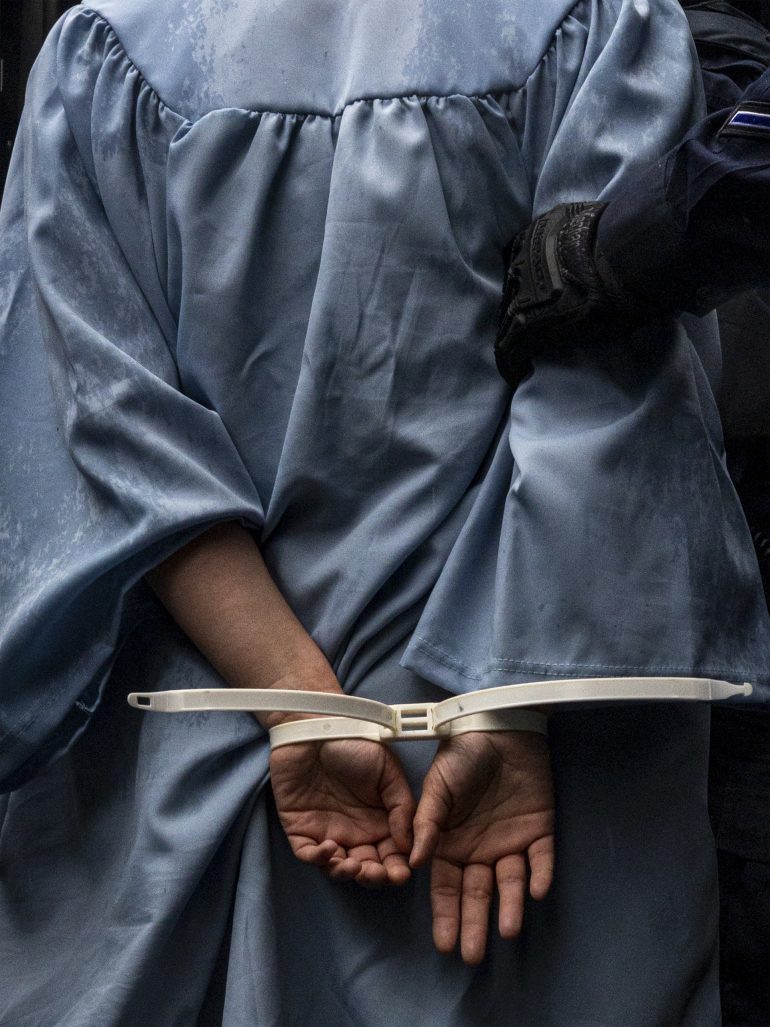 Person in a blue graduation gown is handcuffed with zip ties and held by a gloved officer.