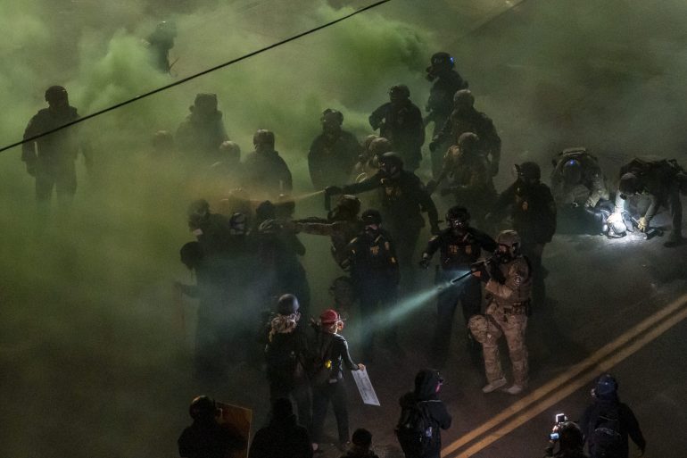 Police in riot gear confront protesters in a street filled with green smoke at night.