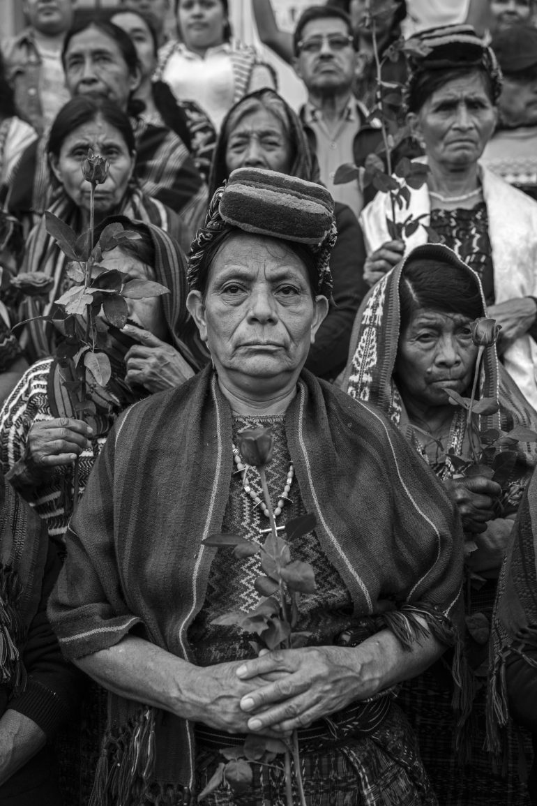 A group of women in traditional clothing hold roses, with one woman standing at the center, facing forward.