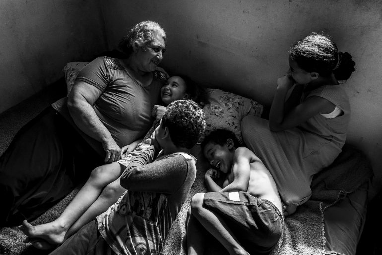 An older woman and four children lie on a bed, smiling and laughing together in a sunlit room.