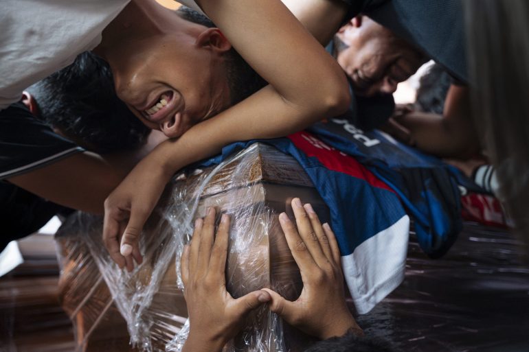 People grieving and crying while embracing a plastic-wrapped casket, with hands touching its surface.