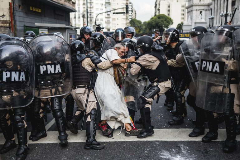 A man in white is pushed by riot police with shields during a protest on a city street.