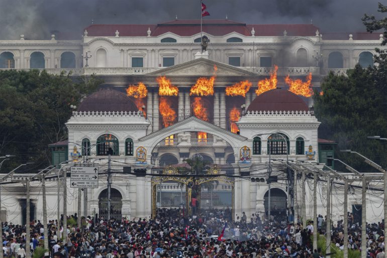 A large crowd gathers as flames engulf the upper floors of a grand white building with domed towers.