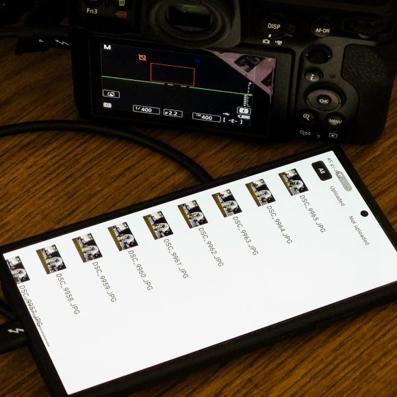 A smartphone and digital camera connected by a cable, displaying photo files on the phone screen.