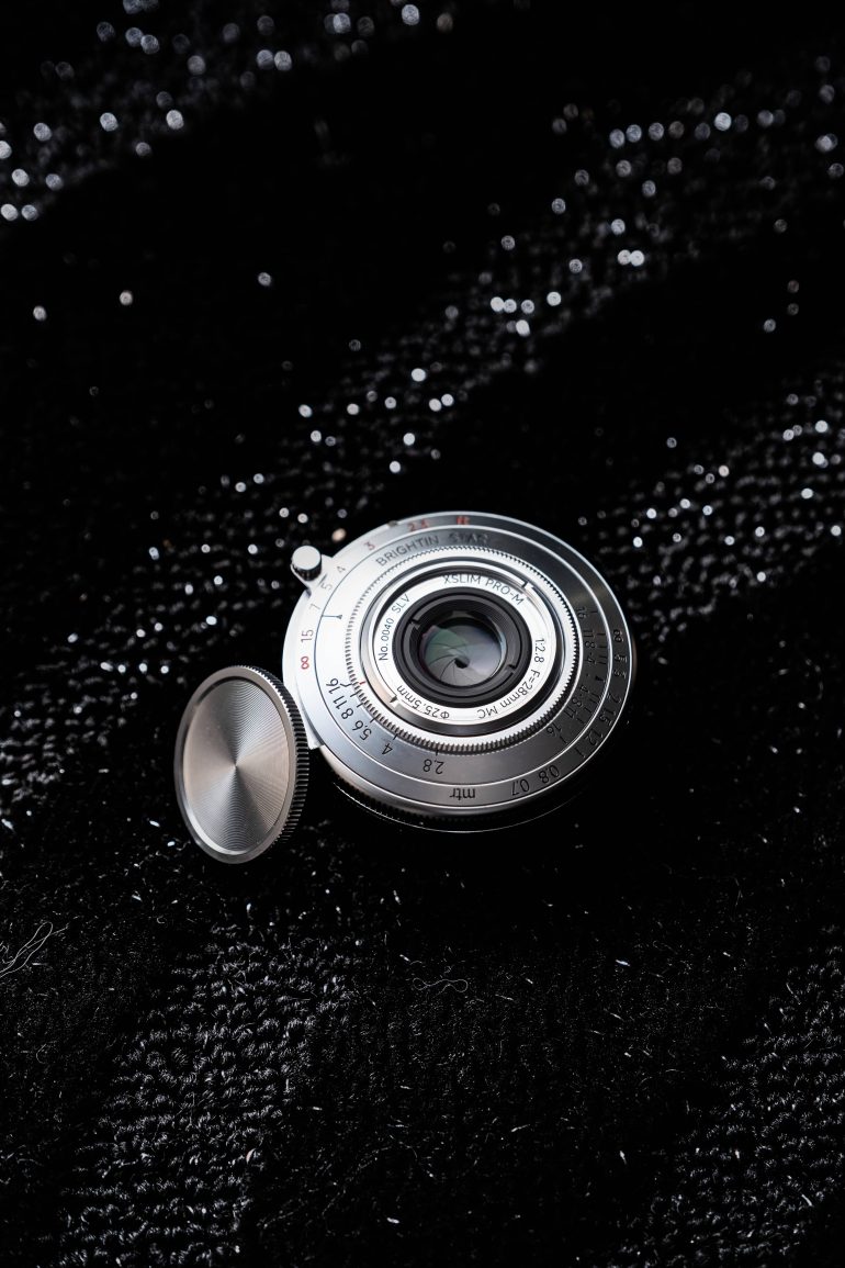 A silver camera lens with its cap off, placed on a textured black fabric background.