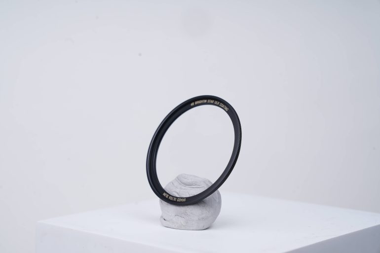 A black circular camera lens filter balanced upright on a small gray stone against a plain white background.