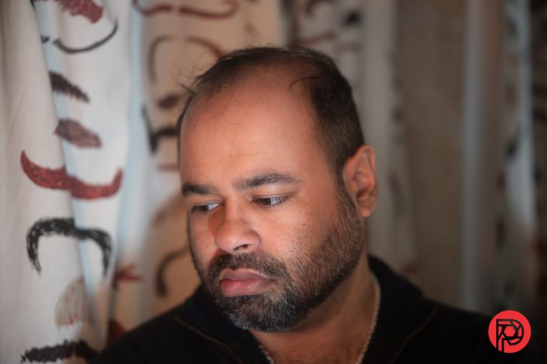 A man with a beard looks down pensively, standing near patterned curtains.