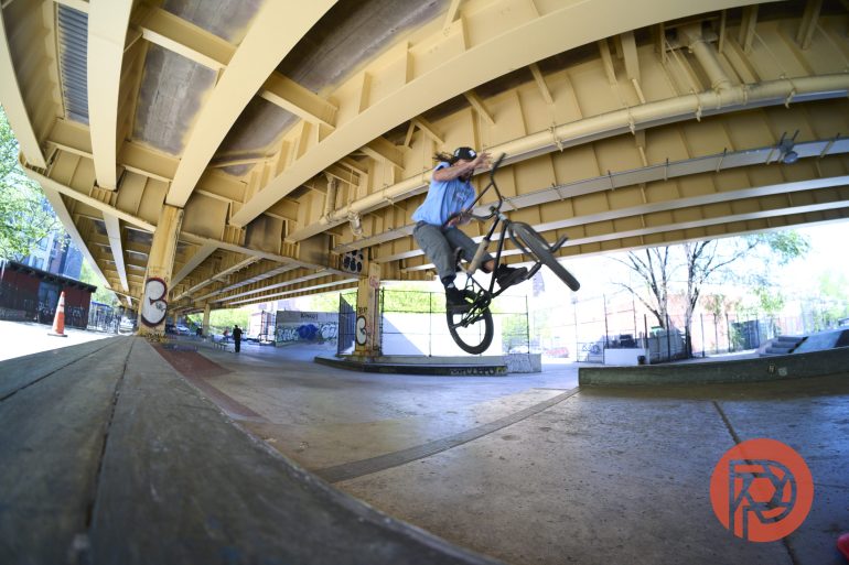 A person performs a trick on a BMX bike under a yellow bridge at an urban skatepark.