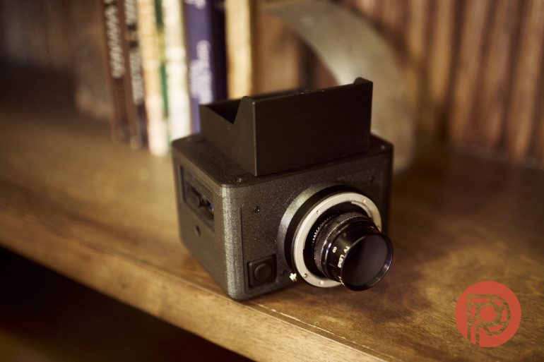 A black vintage-style camera sits on a wooden shelf near some books.