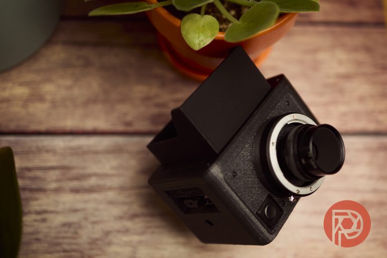 Black vintage camera on a wooden surface next to a potted plant; red logo in the bottom right corner.
