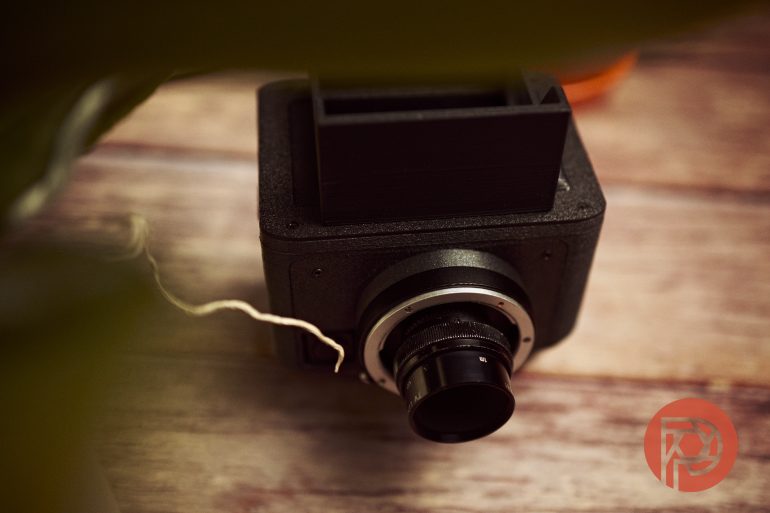A black box camera with a protruding lens sits on a wooden surface, partly obscured by green leaves.