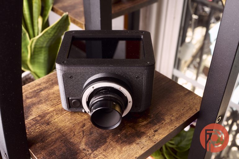 A black digital camera with a large lens sits on a wooden shelf near green plants and a window.