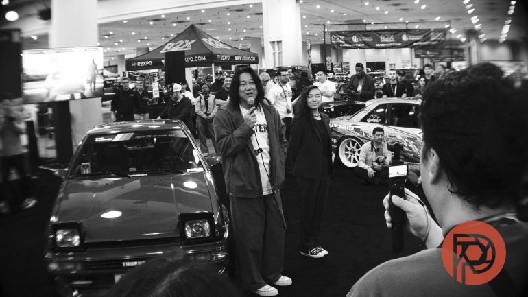 Two people stand by a car at an indoor auto show while a crowd takes photos and watches.