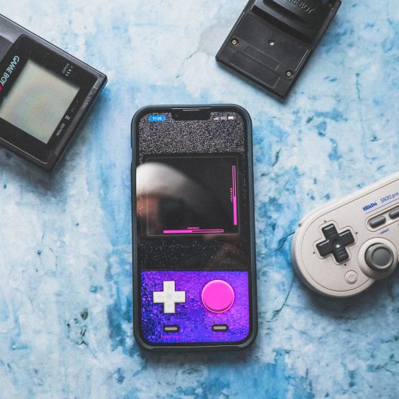 A smartphone with a retro gamepad case, a game controller, and two handheld consoles on a blue surface.
